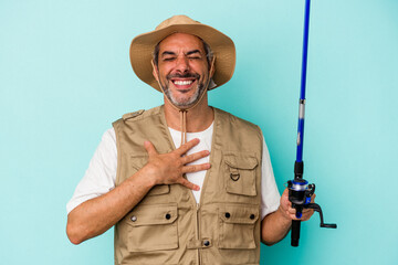 Middle age caucasian fisherman holding rod isolated on blue background  laughs out loudly keeping hand on chest.