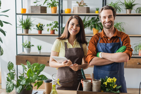 Positive florists in aprons with digital tablet looking at camera near plants and coffee in flower shop