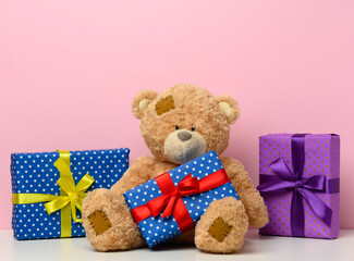 cute brown teddy bear holding a box wrapped in paper and red silk ribbon on white table. Prize and congratulations, pink background
