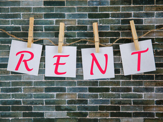Text rent Note is written on a white square paper sheets that hangs with a clothespin on a rope on grey brick wall background