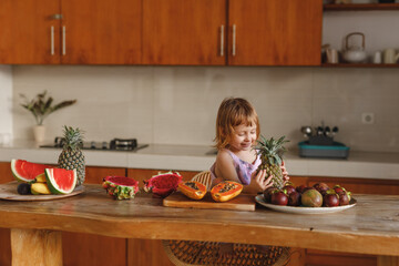 Healthy food. happy child girl and a lot of fruit. Cute little girl holding pineapple while sitting nar wooden table in home kitchen