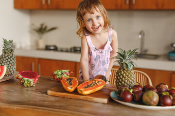 Child healthy food and balanced nutrition.  Fruit breakfast. Smiling little girl in the kitchen ready to have tropical fruits - pineapple, papaya, mangosteen, watermelom