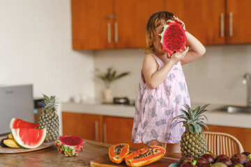 Cute little girl child having fun on kitchen with exotic tropical fruits, cover her head with big pitahaya dragon fruit. Beautiful little girl having breakfast with fruits