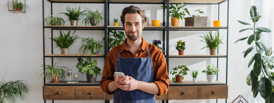 Florist holding smartphone and looking at camera in flower shop, banner