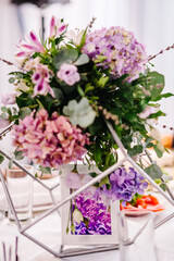 Festive table decorated with composition of violet, purple, pink flowers and greenery in the banquet hall. Table newlyweds in the area on wedding party.