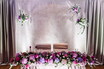 Festive table, arch decorated with composition of violet, purple, pink flowers and greenery in the banquet hall. Table newlyweds in the area on wedding party.