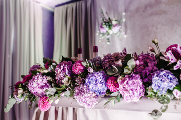 Festive table decorated with composition of violet, purple, pink flowers and greenery in the banquet hall. Table newlyweds in the area on wedding party.