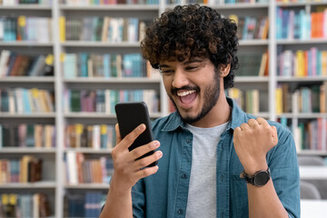 Excited student is happy holding mobile phone and cheering standing in library
