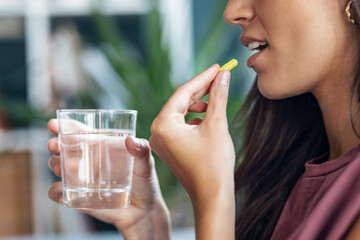 Young woman taking pills while holding glass of water sitting on the couch at home.