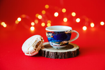 A cup of espresso coffee on a wooden stand and a bitten macaroon on a red background and against the background of garland lights, close-up. Christmas breakfast.