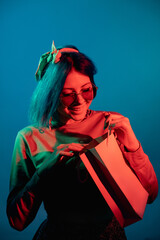 Giveaway offer. Shopping bonus. Gift surprise. Cheerful woman in red neon light with paper bag present isolated on blue background.