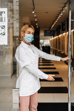 Young And Attractive Female Optometrist With Face Protective Mask Standing At Open Optical Store Doors And Looking Outside. She Is Confident And Serious. Covid-19 Open For Business Concept.