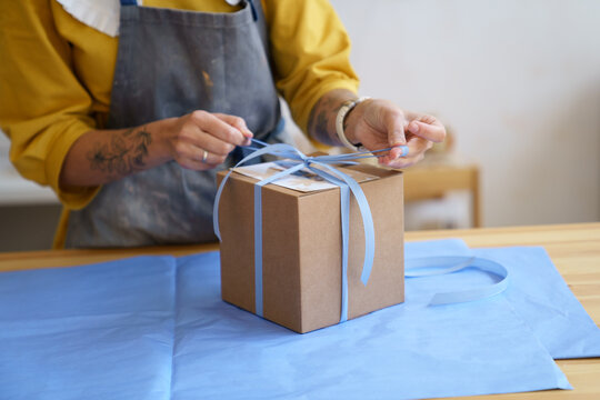 Female ceramist in apron packing gift box with craft crockery for delivery. Closeup of female artist prepare present for shipping to online order customer. Small business and entrepreneurship concept