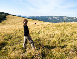 Rear view of a hiker woman dressed in a black hoodie standing on a grassy field in the mountains. Hot summer day in the mountains. Smiling female face.