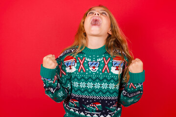 brunette kid girl in knitted sweater Christmas over red background  looks with excitement up, keeps hands raised, notices something unexpected.