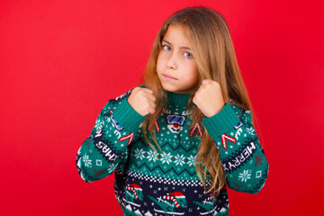 Portrait of attractive brunette kid girl in knitted sweater christmas over red background holding hands in front of him in boxing position going to fight.