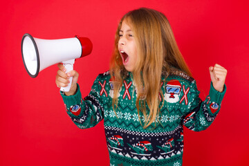 brunette kid girl in knitted sweater christmas over red background communicates shouting loud holding a megaphone, expressing success and positive concept, idea for marketing or sales.