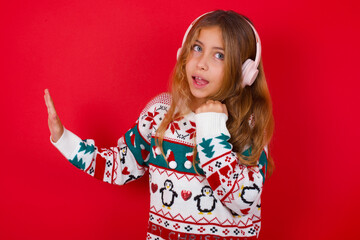 Happy brunette kid girl in knitted sweater christmas over red background sings favourite song keeps hand near mouth as if microphone wears wireless headphones, listens music