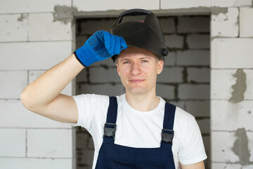 Male construction worker wearing a welding helmet and blue gloves against a brick wall.