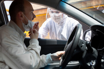 A man wearing a protective suit, gloves, surgical mask, and face mask is testing the coronavirus co id-19 on another man sitting in a car.