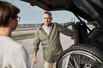 Portrait of mature couple unloading wheelchair out of car trunk in parking lot outdoors, copy space - Powered by Adobe