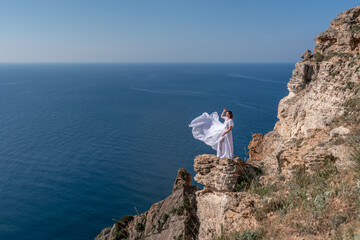 A beautiful young woman in a white light dress with long legs stands on the edge of a cliff above the sea waving a white long dress, against the background of the blue sky and the sea.