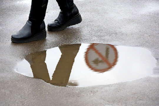 Rain in city, woman in boots walks around the puddle on city street. Road sign forbidding a turn are reflected in water, concept of choosing the wrong way