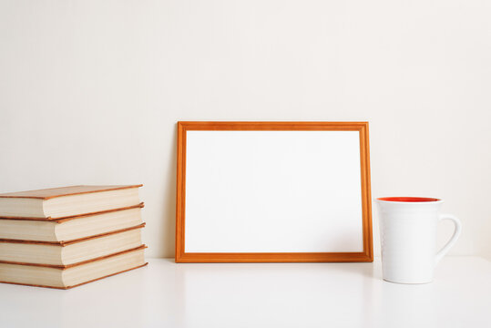Horizontal frame mock-up with empty space for branding, picture, photo or text. Minimalistic interior template, blank frame, stack of books and cup of coffee or tea on table indoors