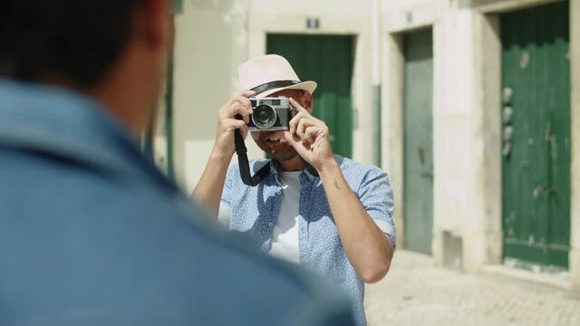 Front view of smiling man taking photo of boyfriend on camera. Happy male person in hat standing in street, taking picture of lover during summer vacation in Europe. LGBT, photoshoot concept