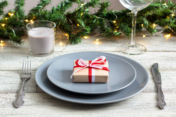 Christmas table setting with gift on grey plate on white table. Holiday view with christmas tree