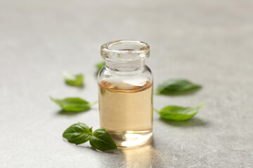 Glass bottle of basil essential oil and leaves on light grey table, closeup