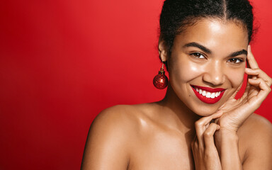 Fashion girl with red lips and beautiful smile, standing in christmas earrings and looking flirty at camera, coquettish smiling face. Black woman posing against red background - obrazy, fototapety, plakaty