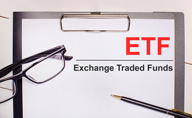 On a light wooden background glasses, a pen and a sheet of paper with the text ETF Exchange Traded Funds. Business concept