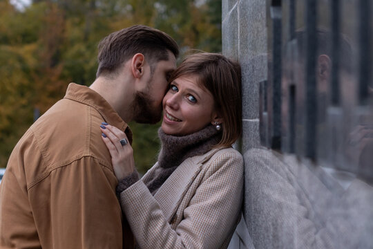 Young man kisses his beloved outside. She hugs him by the shoulders and smiles at the camera. Side view image