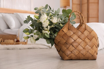 Stylish wicker basket with fresh eucalyptus branches and flowers on floor in bedroom