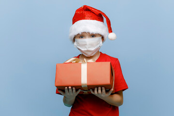 Portrait of a happy little boy in a medical mask, wearing a Christmas hat, holding a gift box and looking into the camera, isolated on a blue background. Copy space