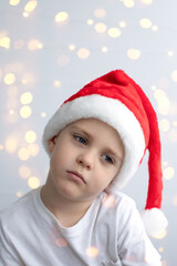A sad little boy in a Christmas red cap on a light festive background with bokeh.