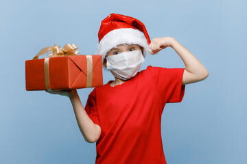 Portrait of a smiling cute boy dressed in a red suit and glasses in a medical mask, holding a gift box against a blue wall