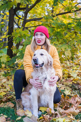 A woman in warm clothes with her retriever is looking somewhere into the distance and sitting near a maple tree among yellow  leaves