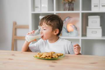 A six-year-old boy is drinking milk with a plate full of cookies in front of him, wearing a white T-shirt. Home cooking, baby food.