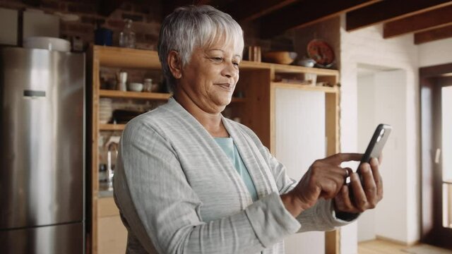 Retired multi-ethnic elderly woman messaging on cellphone. Standing in modern kitchen, happy