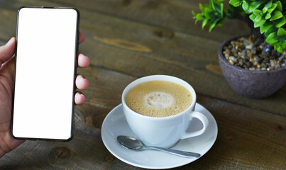 Top view mockup image of a man holding mobile phone with blank white desktop screen while drinking coffee on wooden table