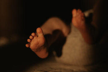 A close-up photo of the foot of a newborn baby in a bodysuit who is lying on a sofa in the sunlight in the evening.