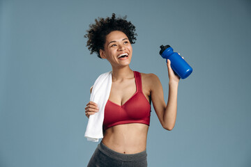 Pensive happy female runner looking up and laughing while drinking water at the studio with blue walls