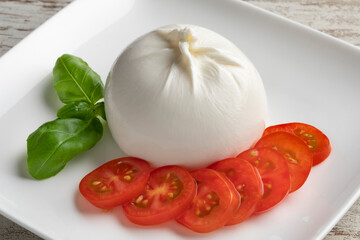 Fresh piece burrata cheese close up on a white dish with tomato slices and basil leaves 