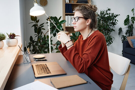 Young woman in eyeglasses sitting at desk with laptop and thinking over her online work at home