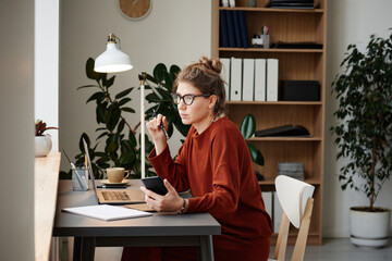 Young woman sitting at the table with laptop and using mobile phone at her work at office