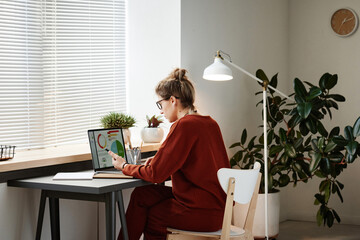 Rear view of woman sitting at the table with laptop and working with financial graphs online at office
