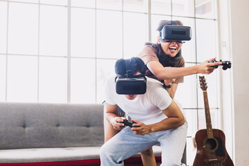 Fototapeta na wymiar Young Asian couple enjoying video games with VR head sets at home while sitting on sofa. Cheerful couple having fun with virtual reality glasses in living room.
