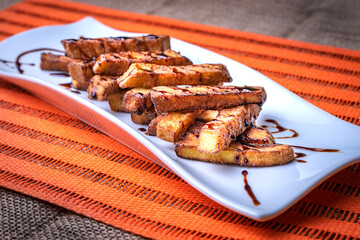 Presentation in rectangular plate of aubergines cut into strips and fried, with sugar cane honey scattered on top, on an orange background.